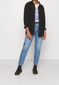 Camisa negra de botones oversize, camiseta gráfica blanca, jeans rectos de mezclilla azul claros, botas negras con cordones y suelas gruesas.