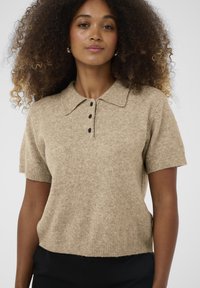 Beige kötött pólóing rövid ujjal, galléros nyakkal és három fekete gombbal, puha textúrával és bordás szegéllyel.