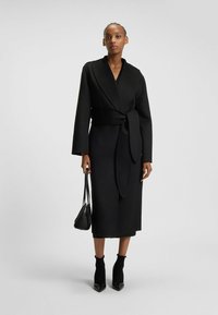 Black wrap coat made of textured fabric, with wide sleeves and a tied waist. Paired with a small black handbag and pointed ankle boots.