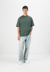 Mørkegrøn bomuld t-shirt med korte ærmer, prydet med tekstgrafik på fronten. Bæres med lyseblå løse jeans og hvide sneakers.