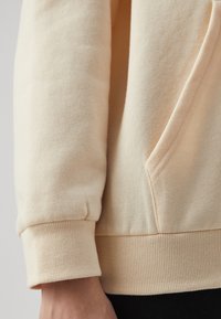 Acercamiento a una manga de sudadera beige con puño de canalé y costura del bolsillo, llevada sobre pantalones negros.