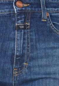 Jeans de mezclilla azul oscuro con etiqueta ajustada, que presentan costuras amarillas contrastantes, un botón de metal y un diseño clásico de cinco bolsillos.