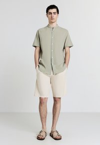 Hellgrünes Kurzarmhemd mit strukturiertem Finish, Frontknöpfen, kombiniert mit beigen Shorts und braunen Sandalen. Einfaches Design, keine Muster.