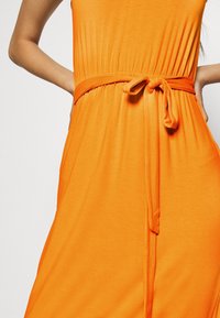 Femme portant une robe sans manches orange avec une ceinture nouée à la taille, les mains placées sur les hanches contre un fond clair.