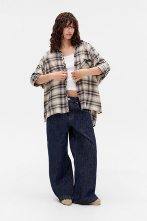 Mlada žena nosi oversized bež i crno kariranu košulju preko bijelog crop topa, široke tamnoplave traperice i svijetle cipele, stojeći na bijeloj pozadini.