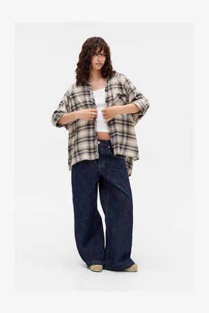 Young woman wearing oversized beige and black plaid shirt over white crop top, wide dark blue jeans, and light-colored shoes, standing on white background.