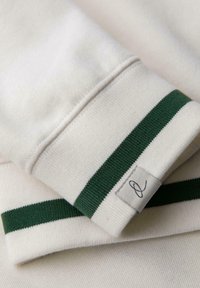 Tejido beige claro con puños acanalados de rayas verdes y una pequeña etiqueta blanca que presenta un logo abstracto negro.