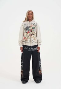 Beżowa bluza z zamkiem i kolorowym nadrukiem graficznym, zestawiona z czarnymi szerokimi spodniami dżinsowymi z logo i ozdobnymi wzorami, noszona z czarnymi butami.