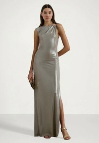 Uma mulher vestindo um vestido longo metálico prateado sem mangas, com uma fenda alta até a coxa, segurando uma pequena clutch e usando saltos dourados.