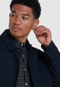 Veste navy avec une fermeture à bouton sur une chemise à carreaux verte et noire. Gros plan sur une silhouette masculine touchant son oreille. Texture lisse.