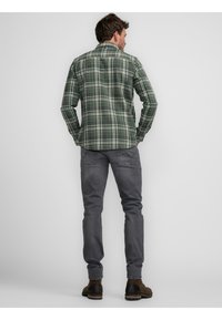 Camisa de cuadros verde y negra de manga larga, con collar de botón, y jeans de mezclilla grises. Botas marrones con una base en contraste.
