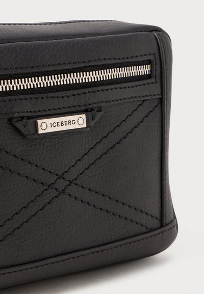 Coin de sac en cuir noir avec une fermeture éclair argentée, des motifs cousus en diagonale et une étiquette de marque en métal portant le nom "ICEBERG" sur une surface texturée.