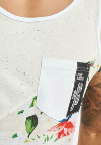 Hvid tanktop med flettet struktur, der har et farverigt blomsterprint og en hvid brystlomme med et sort logo mærke.