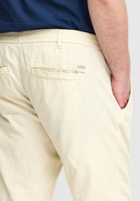 Lys beige bukser med struktureret stof, elastisk talje og to baglommer. Den ene lomme har et lille mærke.
