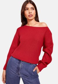 Pulover roșu cu model ribat, cu umerii descoperiți și mâneci largi, dintr-un material tricotat texturat, asociat cu blugi albaștri cu talie înaltă.