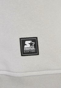 Logo patch Starter Black Label sur un tissu tricoté gris clair avec couture visible et bord côtelé en bas.