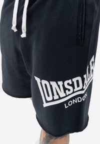 Mørkeblå shorts lavet af bomuld med hvide snore og et stort hvidt "LONSDALE" logo på venstre ben. Har en blød tekstur.
