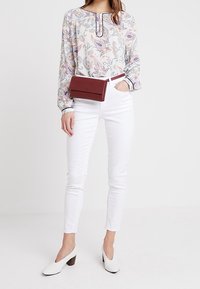 Vit långärmad blus med färgglad paisleymönster, tillsammans med vita skinny jeans, en vinröd bältväska och vita klackskor.