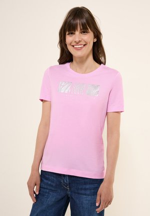 T-shirt di cotone rosa con maniche corte e scollatura rotonda. Presenta un design grafico di testo argentato sul davanti. Indossato con jeans in denim blu.