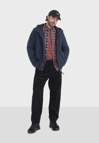 Námornícka kapucňová bunda cez červenú kockovanú košeľu, kombinovaná s čiernymi kordovým nohavicami a čiernymi teniskami. Neformálny outfit so vrstvenými textúrami.