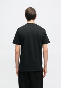 Άτομο με κοντά καστανά μαλλιά, ντυμένο με ένα απλό μαύρο μπλουζάκι και μαύρο παντελόνι, που στέκεται μπροστά σε λευκό φόντο.