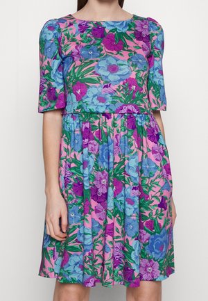 Woman wearing a short-sleeve, knee-length dress with bright pink, blue, and purple floral pattern and green leaves on a light pink background.