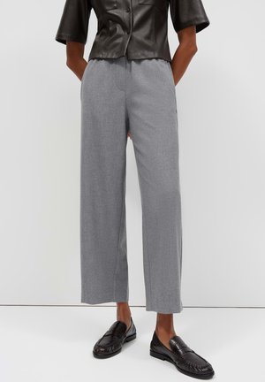 Persoon draagt grijze wijde broek, zwart leren kortärmelig overhemd en zwarte leren loafers, staand tegen een witte achtergrond.