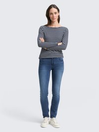 Námornícký a bílý pruhovaný top s dlouhým rukávem, přiléhavý střih, spárovaný s modrými skinny džíny a krémovými teniskami, stojící s překříženýma rukama.