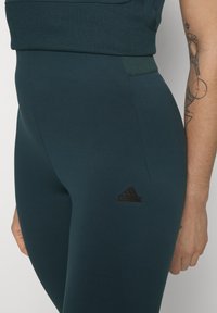 Leggings teal cu talie înaltă, fabricate dintr-un material elastic, cu un talie ribbed și un mic logo Adidas negru aproape de gleznă.
