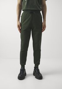 Zelené tepláky s elastickým pasem a stahovací šňůrkou, s černými proužky po stranách a zúženými nohavicemi. Nošeny s černými masivními teniskami.