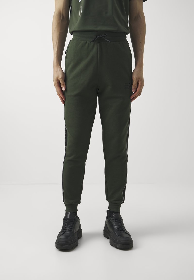 Zelené tepláky s elastickým pasem a stahovací šňůrkou, s černými proužky po stranách a zúženými nohavicemi. Nošeny s černými masivními teniskami.