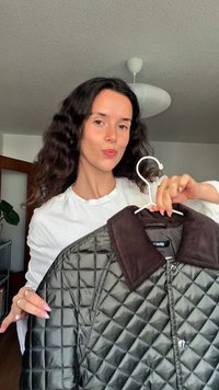 Γυναίκα με μακριά σκούρα μαλλιά που φοράει λευκό πουκάμισο κρατώντας ένα σκουρό πράσινο τζάκετ με καφέ γιακά σε άσπρη κρεμάστρα μέσα σε εσωτερικό χώρο.