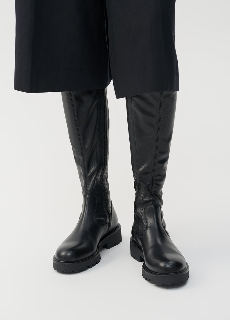 Stivali neri in pelle al ginocchio con una texture liscia, suole spesse e cerniere laterali. Il design presenta una sottile lavorazione delle cuciture.