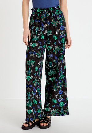 Kvinde iført sorte bukser med vidde, med blå og grøn tropisk blomster- og fugleprint, kombineret med sorte sandaler og en blå top.