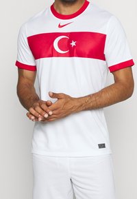 White athletic shirt with red horizontal band featuring a crescent moon and star, red collar and sleeves, Nike logo at the chest.