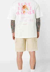 T-shirt in cotone bianco con grafica rosa: "AMORE," "AMORE ESTIVO," e un'immagine. Abbinare a pantaloni cargo beige con tasche laterali.