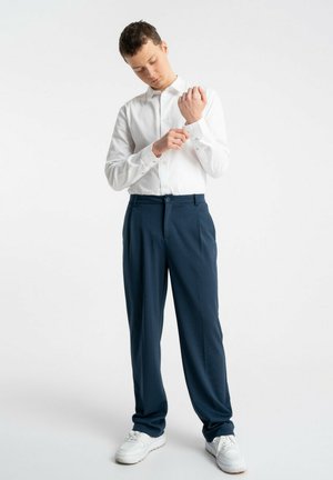 Młody mężczyzna w białej koszuli zapiętej na guziki, poprawiający mankiet, ubrany w granatowe spodnie i białe trampki, stojący na jednolitym tle.