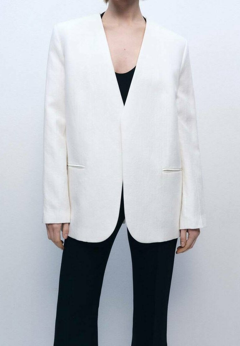 Personne portant un blazer blanc sans col sur un top noir et un pantalon noir, debout devant un fond clair uni.