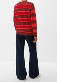 Gestreifter Pullover in Rot- und Brauntönen mit rundem Ausschnitt, kombiniert mit dunklen, weiten Jeans und weißen Sneakers. Hat gerippte Bündchen und Saum.