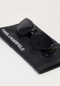 Sorte firkantede solbriller med mørke linser og en blank ramme, som hviler på et sort læderetui med hvid "KARL LAGERFELD" tekst.