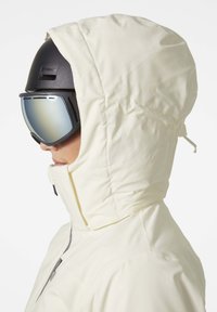 Witte skijas met een aansluitende capuchon, ritssluiting en een hoge kraag. Draagt een zwarte helm en reflecterende goggles.