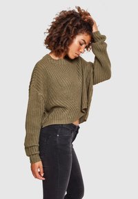 Pull en maille vert olive au design court, avec des poignets côtelés et une couture latérale ouverte, associé à un jean noir ajusté.