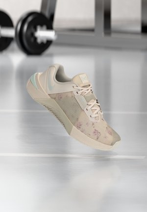 Light beige athletic shoe with pastel blue accents and floral pattern, floating above a reflective gym floor with blurred weights in background.
