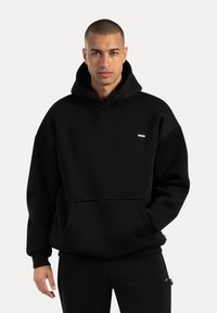 Mann in schwarzem, oversized Hoodie und passender Hose, steht mit einer Hand in der Tasche vor einem schlichten Hintergrund.