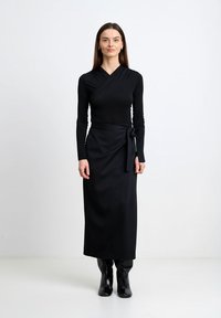 Kobieta stojąca na białym tle, ubrana w czarną bluzkę z kopertowym dekoltem, midi kopertową spódnicę zawiązaną w talii oraz czarne kozaki za kolano.