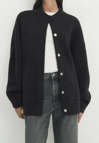 Donkergrijze gebreide cardigan met een hoge kraag, voorzakken en zilveren knopen. Heeft een ontspannen pasvorm en een zachte textuur.