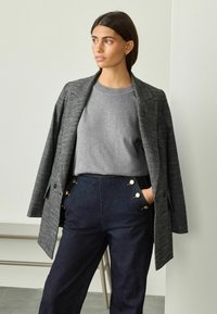 Grå rutigt kavaj över en grå tröja och mörkblå midjehöga byxor med gulddetaljer på knapparna, vilket skapar en lager-på-lager, strukturerad look.