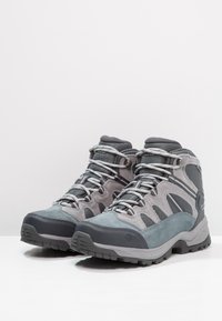 Scarpe da trekking grigie realizzate con una combinazione di suede e materiali in rete, caratterizzate da una caviglia alta, superficie texturizzata e suole in gomma durevoli con trazione.