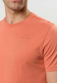 Homme portant un T-shirt à manches courtes couleur corail avec un petit logo brodé représentant deux personnages assis dos à dos sur la poitrine.
