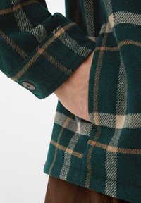 Camicia flanella a quadri verdi con polsini con bottoni, texture spazzolata e dettagli in crema e marrone, con una mano appoggiata in tasca.